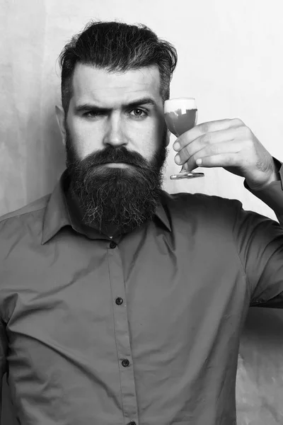 Bearded man, long beard. Brutal caucasian serious hipster with moustache in brown shirt holding alcoholic red shot on golden texture backgroun — Stock Photo, Image