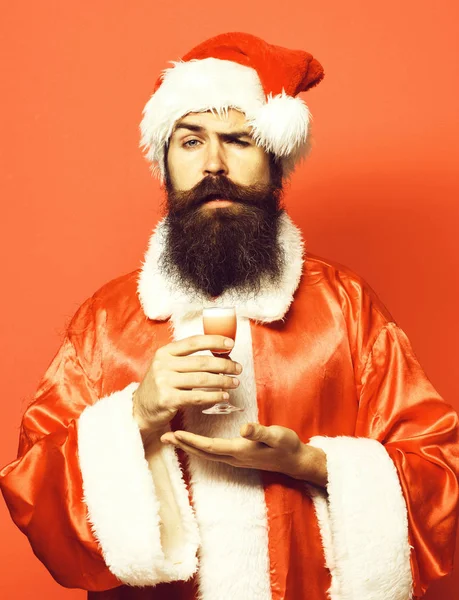 Красивий бородатий Санта Клаус чоловік з довгою бородою на серйозному обличчі тримає келих алкоголю, знятий на руці в різдвяному або різдвяному светрі та новорічному капелюсі на червоному студійному фоні — стокове фото