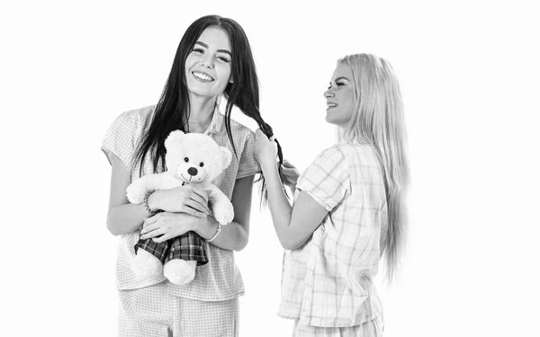 Sisters, best friends in pajamas making braid, hairdo each other. Girls in pink pajamas, isolated white background. Ladies on smiling faces with plush toy bear look cute. Girlish leisure concept — Stock Photo, Image