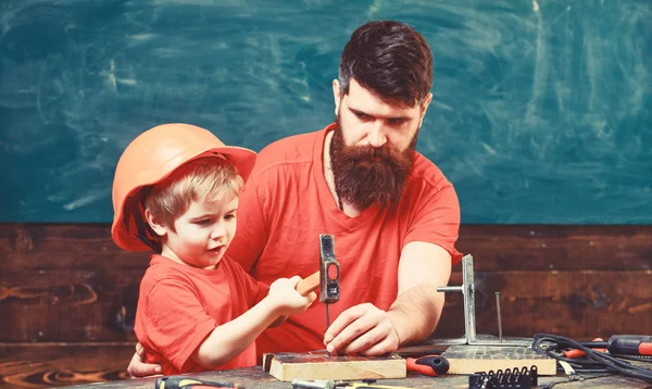 Father with beard teaching little son to use tools, hammering, chalkboard on background. Little assistant concept. Boy, child busy in protective helmet learning to use hammer with da