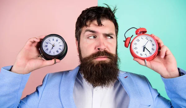 人留胡子的嬉皮士拿着两个不同的钟。这个不折不扣的家伙困惑地面对着时间变化的问题。更改时区会影响运行状况。时区。改变时钟会影响你的健康吗？ — 图库照片
