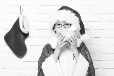 Noel Baba 'nın kırmızı gözlüklü genç ve yeni yıl şapkalı dekoratif noel ya da xmas çoraplı ya da beyaz tuğla duvar arkasında pembe domuz kumbarasını öpen sevimli çocuğu.