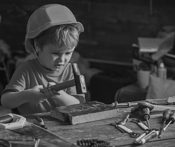 Gyermek sisak aranyos játszik, mint építője, vagy javítóműhely, javítás, vagy handcrafting. Kisgyermek elfoglalt arc játszik otthon a műhelyben. Handcrafting fogalom. Gyerek fiú köröm sulykolja fa tábla — Stock Fotó