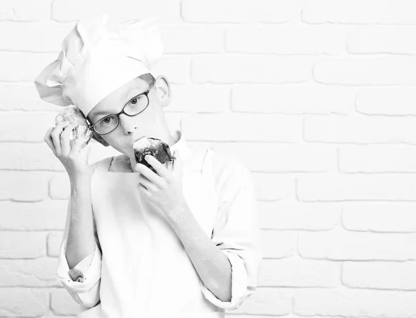 Young boy cute cook chef in white uniform and hat on stained face flour with glasses holding chocolate cakes on brick wall background, copy spac — Stock Photo, Image