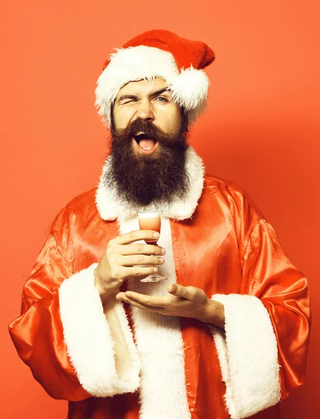 Jóképű szakállas télapó claus férfi hosszú szakállal mosolygós arcon tartó üveg alkoholos lövés kéznél karácsonyi vagy karácsonyi pulóver és szilveszteri kalap piros stúdió háttér — Stock Fotó