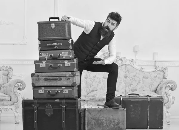 Man, butler with beard and mustache delivers luggage, luxury white interior background. Macho elegant on shocked face standing near pile of vintage suitcase. Luggage and travelling concept