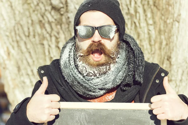 Stilig lycklig man, hipster, med skägg och mustasch i svarta solglasögon täckt med vit frost håller tom svarta tavlan och tummen upp vinterdag på naturlig bakgrund, kopia spac — Stockfoto