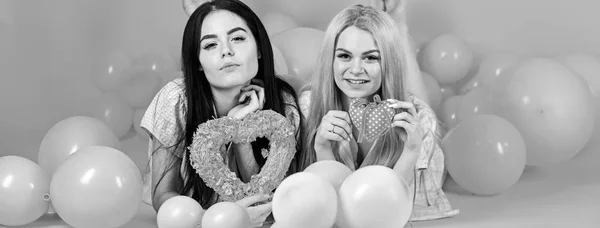 Brunette and blonde cute women in pink pajamas posing on camera with pink hearts while lay near air balloons at slumber party over pink backgroun — Stock Photo, Image