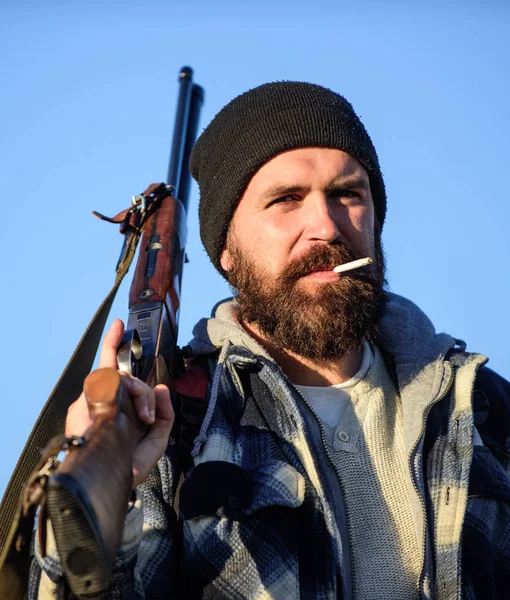 Hunter with rifle gun close up. Guy bearded hunter spend leisure hunting and smoking. Hunting masculine hobby concept. Man brutal bearded guy gamekeeper blue sky background. Brutality and masculinity