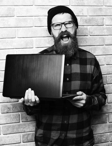 Bearded man, long beard. Brutal caucasian happy unshaven hipster holding laptop in red black checkered shirt with hat and glasses on beige brick wall studio backgroun
