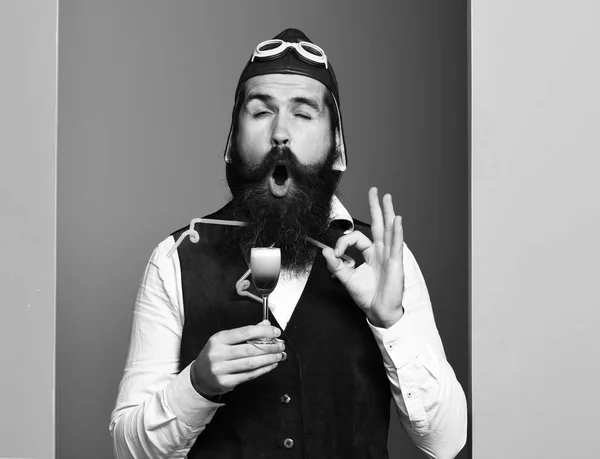 Jóképű szakállas pilóta vagy pilóta férfi hosszú szakállal és bajusszal vicces arc gazdaság üveg alkoholos lövés vintage bőr mellény kalap és szemüveg színes stúdió háttér — Stock Fotó