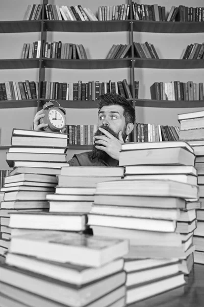 Άνθρωπος, επιστήμονας που κρυφοκοιτάζει από σωρούς από βιβλία με ξυπνητήρι. Δάσκαλος ή μαθητής με τη γενειάδα μελέτη στη βιβλιοθήκη. Προθεσμία έννοια. Ο άνθρωπος στο πρόσωπο έκπληξη που βλέπουν ρολόι, ράφια σε φόντο — Φωτογραφία Αρχείου