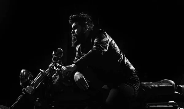 Masculinity concept. Macho, brutal biker in leather jacket riding motorcycle at night time, copy space. Man with beard, biker in leather jacket sitting on motor bike in darkness, black background.