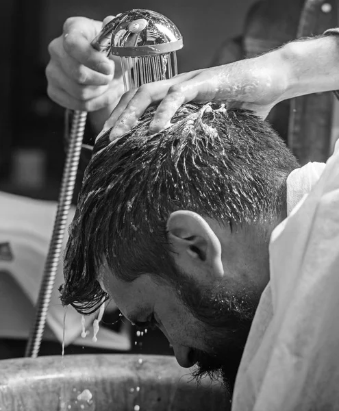 Hombre barbudo cliente de barbería hipster. Los peluqueros lavan los cabellos del hipster barbudo. Concepto de barbería. Hombre con barba y bigote con toalla en los hombros, manos masculinas con ducha en el fondo — Foto de Stock