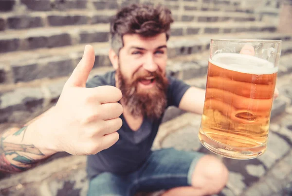 Hipster på glada ansikte dricker öl. Fatöl koncept. Kille med resten med öl, selektivt fokus. Mannen med skägg och mustasch innehar glas med öl och visar tummen upp, stentrappa bakgrund — Stockfoto