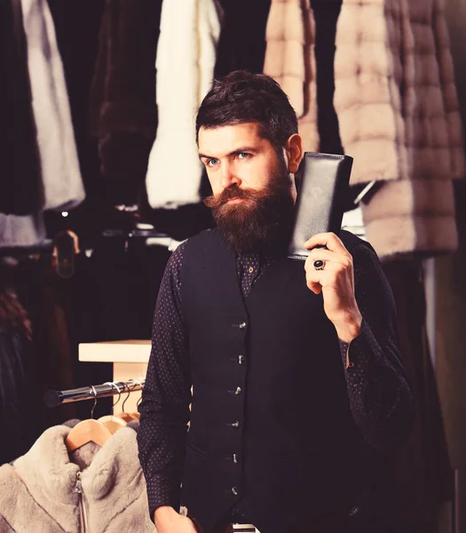 El tipo con la cara pensativa sostiene la cartera. Macho con billetera delante de ropa peluda de lujo. concepto de ropa de lujo. Hombre con barba y bigote en peluquería — Foto de Stock