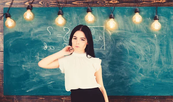 Lady teacher on calm face stands in front of chalkboard. Woman with long hair in white blouse stands in classroom. Elite school concept. Teacher ready to start lesson, copy space