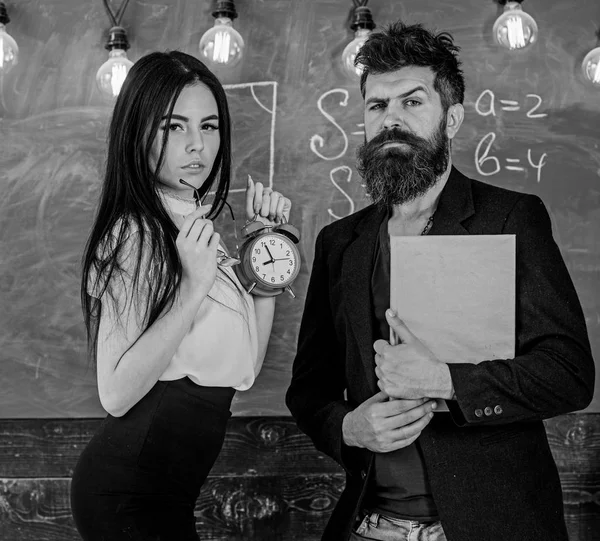 Concepto de disciplina. Hombre con libro de retención de barba y sexy profesora de niña sostiene el reloj despertador, pizarra en el fondo. Profesora y estricta maestra de escuela se preocupan por la disciplina y las reglas en la escuela — Foto de Stock