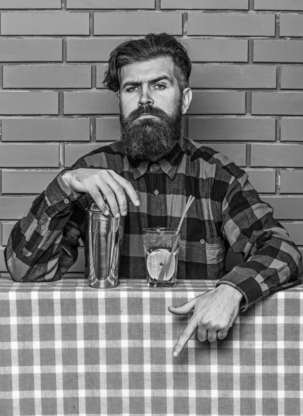 Conceito de menu de bar. Homem de camisa quadriculada perto de shaker e coquetel, fundo da parede de tijolo. O barman recomenda tentar a bebida. Barman com barba no rosto estrito apontando para baixo com o dedo indicador — Fotografia de Stock