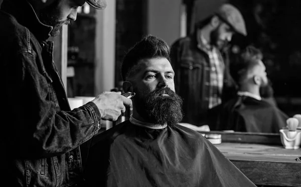 Hipster with beard covered with cape trimming by professional barber in stylish barbershop. Grooming concept. Barber busy with grooming beard of hipster client, mirror reflexion on background — Stock Photo, Image