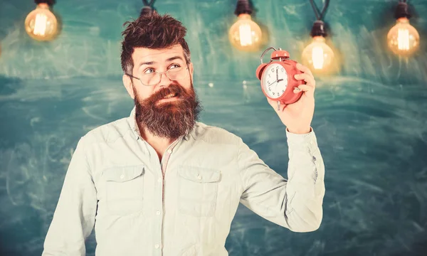 El profesor de anteojos tiene despertador. hipster barbudo sostiene reloj, pizarra en el fondo, espacio de copia. Hombre con barba y bigote en la cara molesta escucha campana de alarma. Concepto de campana escolar — Foto de Stock