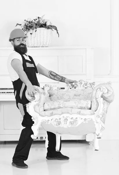 Delivery service concept. Man with beard, worker in overalls and helmet lifts up armchair, white background. Loader carries armchair. Courier delivers furniture in case of move out, relocation