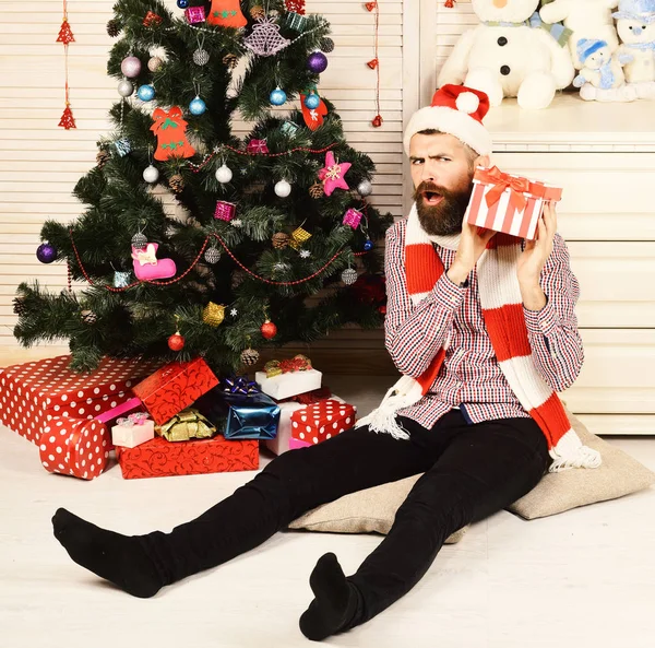 Guy in hat and scarf sits by Christmas tree. — Stock Photo, Image