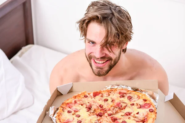 Guy holds pizza box sit bed in bedroom or hotel room. Student is at home on the bed in a bright apartment eating a tasty pizza. Cropped image of shirtless sexy man with pizza on bed. Dinner in bed. — Stock Photo, Image