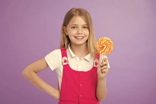 Mädchen lächeln mit Lutscher auf violettem Hintergrund. glückliches Kind mit wirbelndem Karamell auf lila Hintergrund. Ein kleines Kind lächelt mit Bonbons am Stock. Essen und Dessert. süß wie Lutscher. Ernährung und Diät — Stockfoto