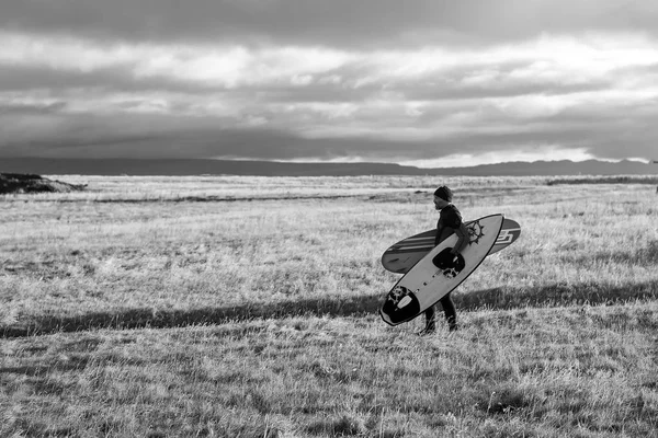 Man surfer walk on grass field in reykjavik, iceland. Man with surfboards on autumn landscape. Sportsman on cloudy grey sky. Sport activity and energy. Vacation and wanderlust — Stock Photo, Image
