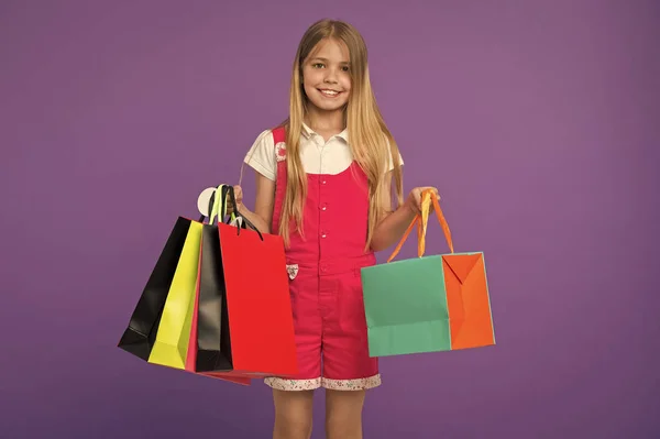 Glada barn med kassar på lila bakgrund. Liten flicka leende med papperspåsar. Kid shopper i mode overall. Holiday förberedelser och firande. Efter dag med shopping. försäljning och svart fredag — Stockfoto