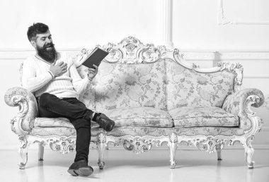 Adam zevk ile eski kitap okuma. Sakal ve bıyık oturur barok tarzı kanepe, tutar kitap, beyaz duvar arka plan üzerinde olan adam. Mizahi edebiyat kavramı. Maço yüz kitap okuma gülüyor üzerinde