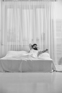 Yatak, döşeme tişörtlü adam arka plan üzerinde beyaz perde TV izlemek. Adam anahtarı kanal için uzaktan kumanda ile ciddi bir yüz. Uzaktan kumanda kavramı. Maço sakallı denetleyicisi elinde tutar