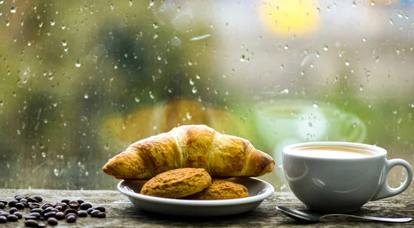 Profiter du café le jour de pluie. Heure du café le jour de pluie. Café frais brassé dans une tasse blanche ou une tasse sur le rebord de la fenêtre. Fenêtre en verre humide et tasse de boisson à la caféine chaude. Boisson café avec dessert croissant — Photo