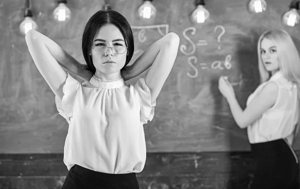 Donne attraenti che si preparano per la lezione. Concetto di studenti e tirocinanti. Ragazza sembra attraente mentre la signora scrive su sfondo lavagna, sfocato. Studente, insegnante in occhiali stare in classe — Foto Stock