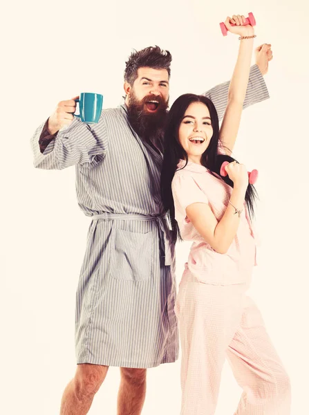 Couple, family on sleepy faces, full of energy. Couple in love in pajama, bathrobe stand isolated on white background. Girl with dumbbell, man with coffee cup. Morning routine concept