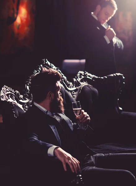 Alcohol, wealthy lifestyle, business, money concept. Boss in luxury suit sits on vintage sofa. Mature man with beard talks to assistant. Bearded man with serious face and champagne glass