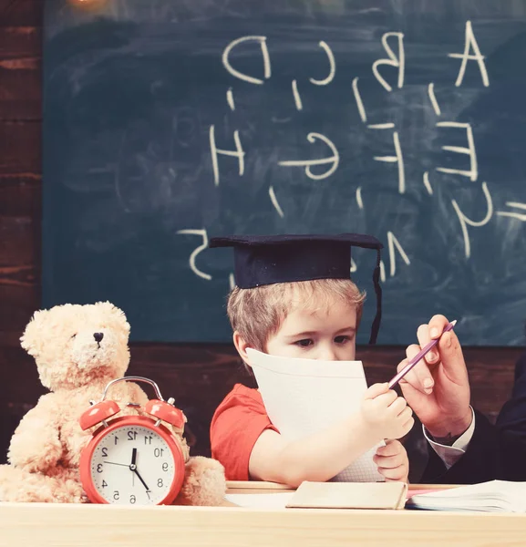 Grundutbildning koncept. Elev i mortarboard, svarta tavlan på bakgrund. Kid studier med häfte, manlig hand ger pennan. Pojken vill skriva eller rita med penna. Barn lära sig att skriva — Stockfoto