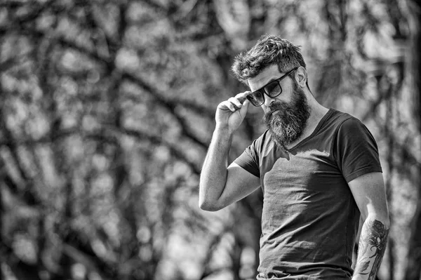 L'uomo barbuto si toglie gli occhiali da sole nella giornata di sole. Concetto di mascolinità. L'uomo con la barba lunga sembra elegante e sicuro di sé. Uomo con barba e baffi sul viso severo, rami sullo sfondo, sfocato — Foto Stock