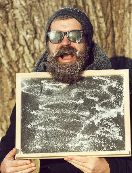 Upphetsad skäggig man, hipster, med skägg och mustasch i svarta solglasögon täckt med vit frost innehar svarta tavlan eller svarta tavlan vinterdag på naturlig bakgrund, kopia spac — Stockfoto