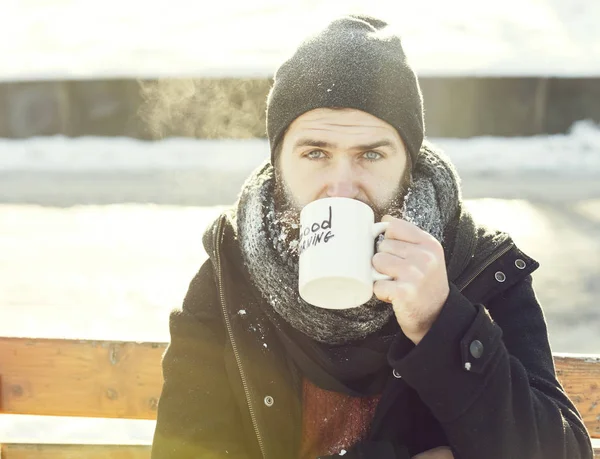 Hombre fruncido, hipster barbudo con barba y bigote cubierto de bebidas heladas blancas de la taza con texto de buena mañana sentado en el banco de madera en el día de invierno nevado al aire libre en el fondo natural —  Fotos de Stock