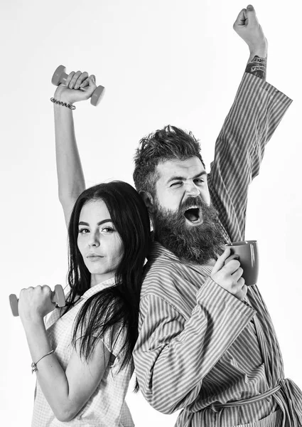 Casal apaixonado no pijama, roupão de banho de costas para trás. Conceito de energia matinal. Menina com haltere, homem com xícara de café, isolado em fundo branco. Casal, família em rostos sonolentos, alongamento — Fotografia de Stock