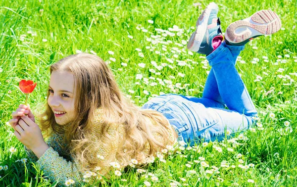 Menina com cabelos longos deitado no gramado, fundo de grama. Criança desfrutar de clima ensolarado primavera enquanto deitado no prado. Menina no rosto sorridente segura flor de tulipa vermelha. Conceito de humor primavera — Fotografia de Stock