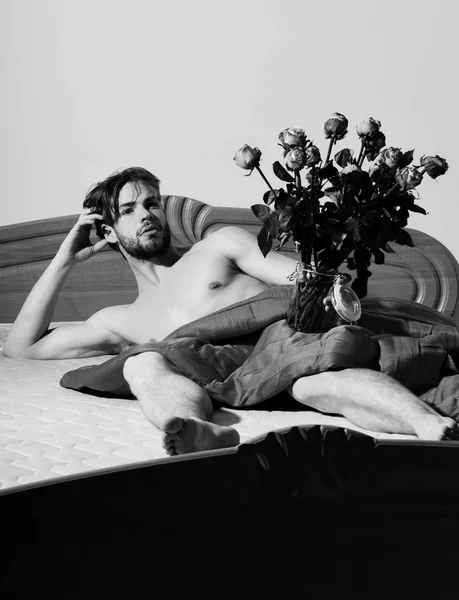 Joven guapo barbudo sexy macho hombre con barba elegante acostado desnudo en la cama debajo de la manta roja sosteniendo florero transparente con ramo de rosas naranjas y muestra abdominales — Foto de Stock