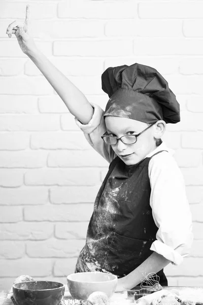 Young boy cute cook chef in red uniform and hat on stained face with glasses standing near table with colorful bowls, tasty cakes, rolling pin and kitchen whisk on white brick wall backgroun — Stock Photo, Image