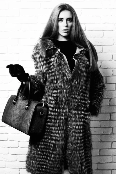 Молодые модные сексуальные довольно богатая женщина с красивыми длинными светлыми волосами в талии пальто серого меха с черными перчатками показывая синий кожаный мешок с отпечатком змеи на кирпичной стене студии фоне — стоковое фото
