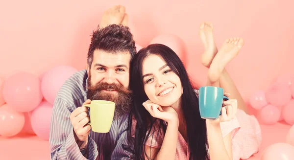 Homem e mulher em rostos sorridentes jazem, fundo rosa. Casal apaixonado bebe café na cama. Casal relaxar de manhã com café. Conceito de tradição familiar. Homem e mulher de roupas domésticas, pijamas — Fotografia de Stock