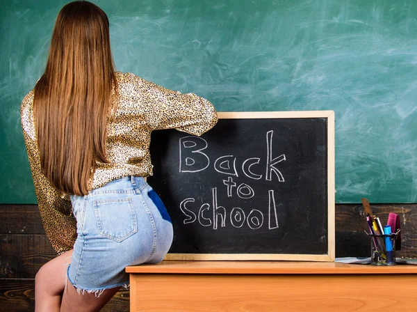 Jupe en jean fille enfreignant les règles de vêtements scolaires. Code vestimentaire scolaire. Étudiant dos et fesses près du tableau. Étudiant professeur mini jupe sexy fesses assis table tableau noir inscription retour à l'école — Photo