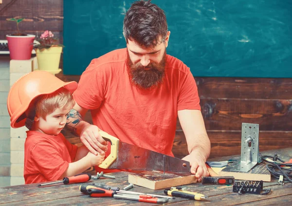 Masculine duties concept. Father, parent with beard teaching little son to sawing with sharp handsaw, carpenter crafts. Boy, child busy in protective helmet learning to use handsaw with dad