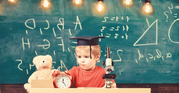 School break concept. Child on concentrated face looks at alarm clock. Pupil waiting for school break. Kid boy in academic cap near microscope, clock and toy in classroom, chalkboard on background